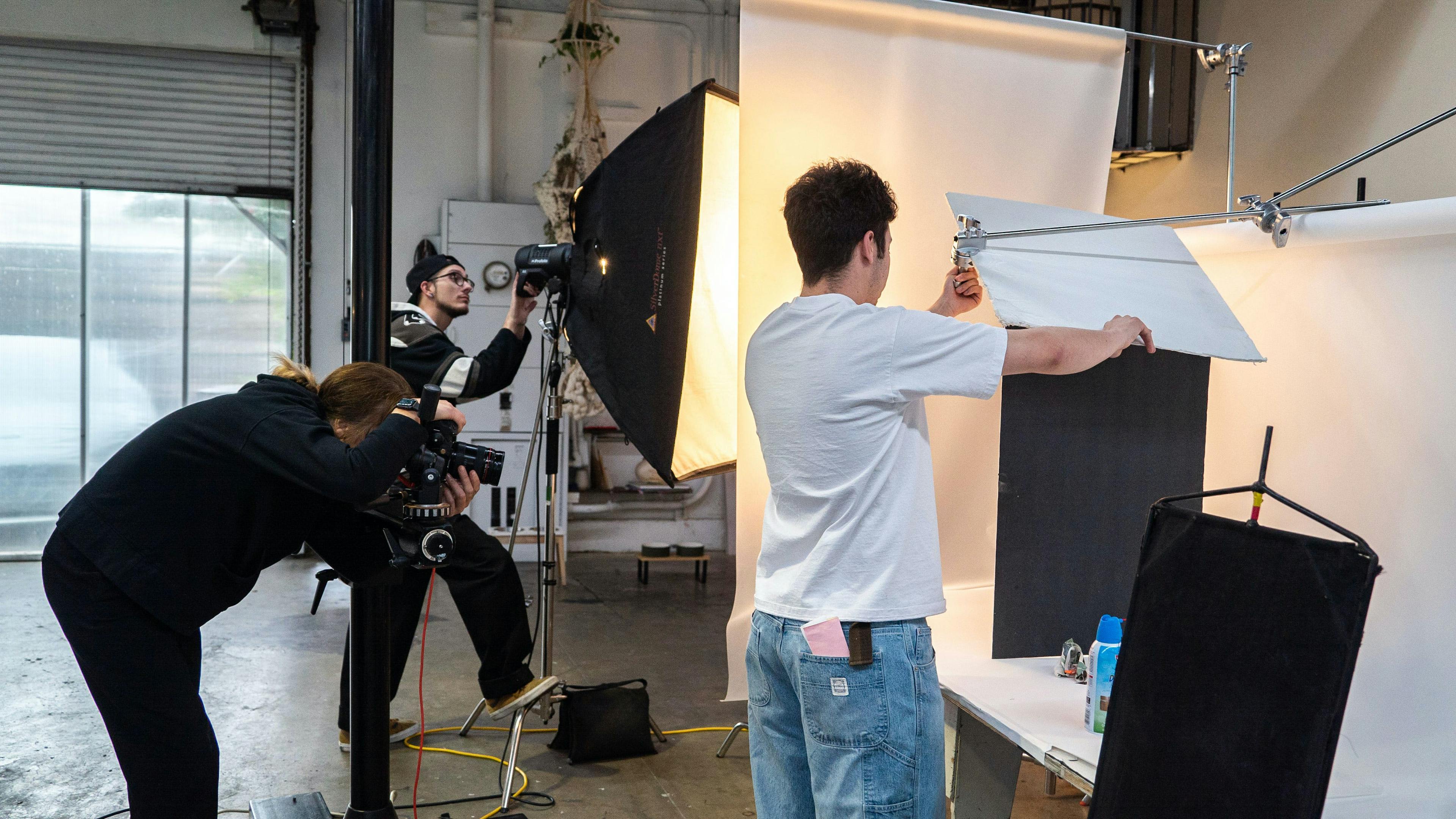 A photography team working on a set with various lighting equipment, cameras, and props. Some team members are adjusting the lighting while others are setting up props and the camera. The atmosphere is busy and focused.
